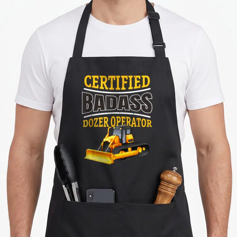 Dozer heavy canvas aprons - Stacked CERTIFIED BADASS DOZER OPERATOR text above yellow cartoon bulldozer on grey — shown on a black cotton canvas apron against a white background