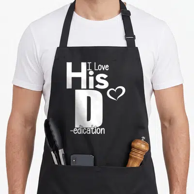 His dedication apron - White vertical text reading I Love His D-edication with a small heart next to the letter D — shown on a black cotton canvas apron against a white background