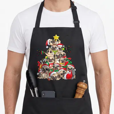 Pug merry christmas apron - Cartoon tan pugs stacked in a pyramid with red hats, yellow star, and green gifts — shown on a black cotton canvas apron against a white background
