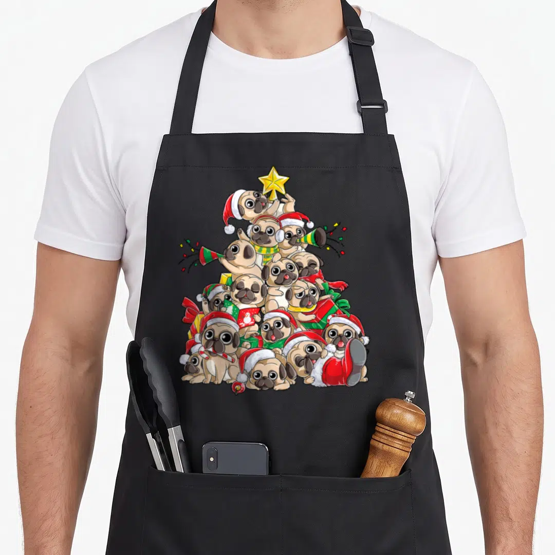 Pug merry christmas apron - Cartoon tan pugs stacked in a pyramid with red hats, yellow star, and green gifts — shown on a black cotton canvas apron against a white background