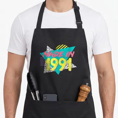 35th birthday apron - Made in 1991 text over cyan triangle with pink yellow letters and black white zigzag patterns — shown on a black cotton canvas apron against a white background