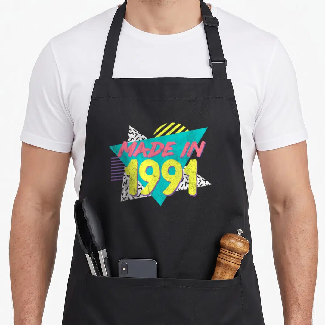 35th birthday apron - Made in 1991 text over cyan triangle with pink yellow letters and black white zigzag patterns — shown on a black cotton canvas apron against a white background