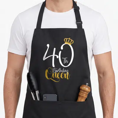 Queen 40th birthday apron - White 40 and gold crown text design on black apron — shown on a black cotton canvas apron against a white background