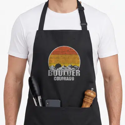 Boulder pride apron - Retro circular sunset with grey mountains and stacked white BOULDER COLORADO text in distressed — shown on a black cotton canvas apron against a white background