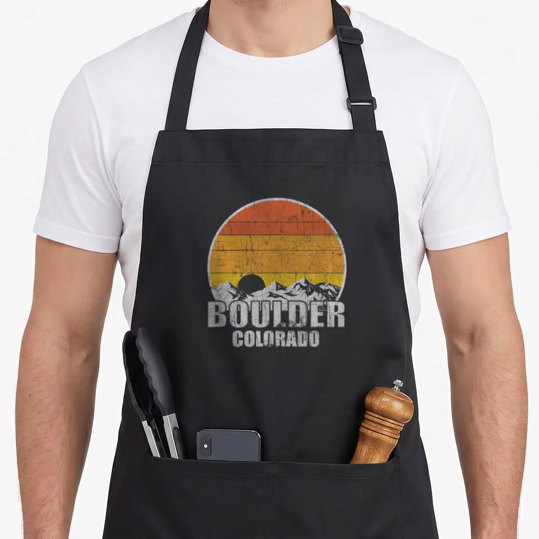 Boulder pride apron - Retro circular sunset with grey mountains and stacked white BOULDER COLORADO text in distressed — shown on a black cotton canvas apron against a white background