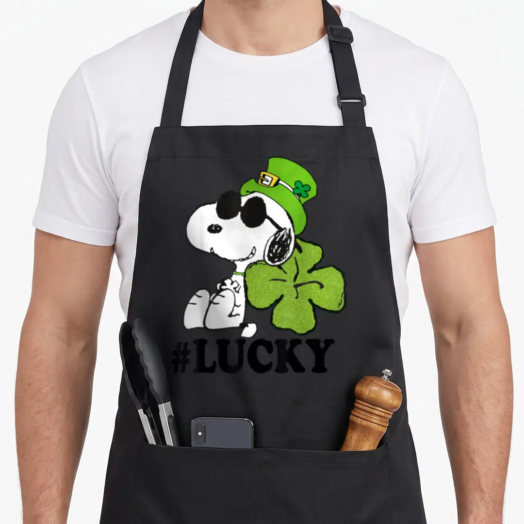 Patrick lucky apron - Snoopy in green hat and sunglasses sitting next to a large green shamrock with #LUCKY text below — shown on a black cotton canvas apron against a white background