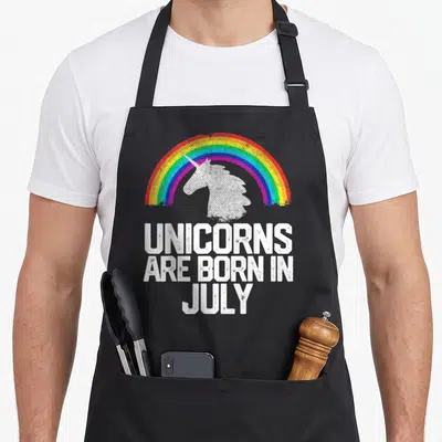 July rainbow apron - White unicorn under rainbow arch with stacked text UNICORNS ARE BORN IN JULY — shown on a black cotton canvas apron against a white background