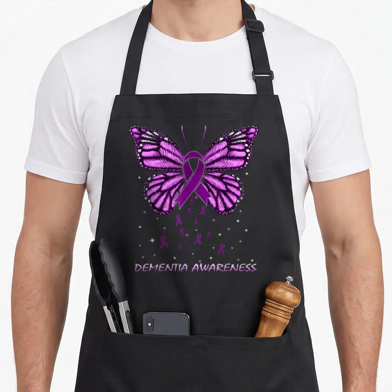 Dementia butterfly apron - Purple butterfly with ribbon body, scattered ribbons, stars, and DEMENTIA AWARENESS text — shown on a black cotton canvas apron against a white background