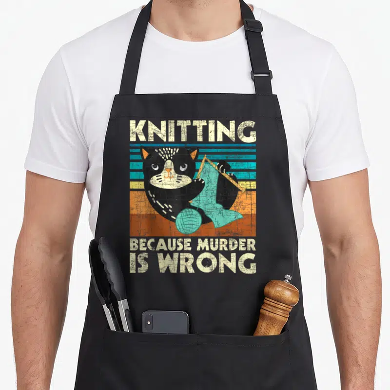 Murder wrong knitting apron - Cartoon black cat knitting teal fabric before colorful stripes with slogan text — shown on a black cotton canvas apron against a white background