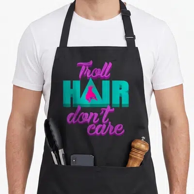 Troll messy aprons - Purple and teal text reading Troll Hair Don't Care with a pink tuft inside the letter — shown on a black cotton canvas apron against a white background