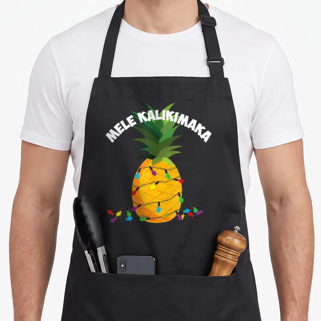Mele pineapple apron - MELE KALIKIMAKA text over cartoon pineapple wrapped in multicolored Christmas lights — shown on a black cotton canvas apron against a white background