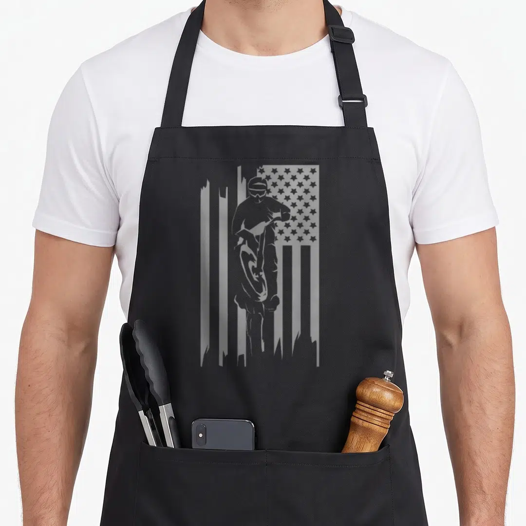 Motocross dirt american flag apron - Grey silhouette of a motocross rider inside a distressed American flag pattern — shown on a black cotton canvas apron against a white background