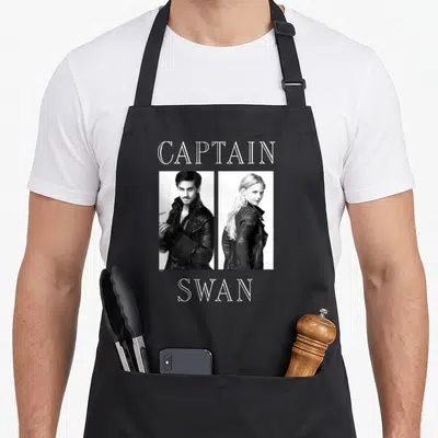 Captain once upon an apron - Two grayscale portraits of a man and woman with CAPTAIN and SWAN text — shown on a black cotton canvas apron against a white background