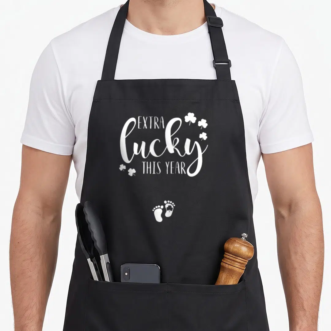 Patty st patrick day aprons - White extra lucky this year text with shamrocks and baby footprints — shown on a black cotton canvas apron against a white background