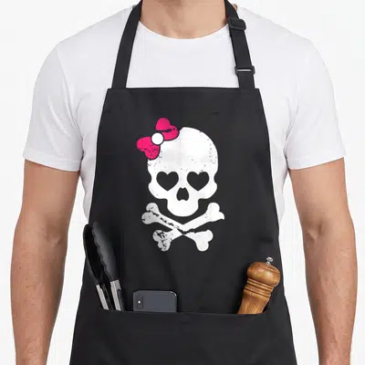 Skull cross apron - White skull with pink bow and heart eyes above two crossed bones — shown on a black cotton canvas apron against a white background