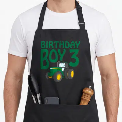 Three farm apron - BIRTHDAY BOY 3 text with green tractor overlapping bottom line — shown on a black cotton canvas apron against a white background