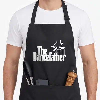 Dancefather dance dad apron - White hands holding puppet control bars above text reading The Dancefather — shown on a black cotton canvas apron against a white background
