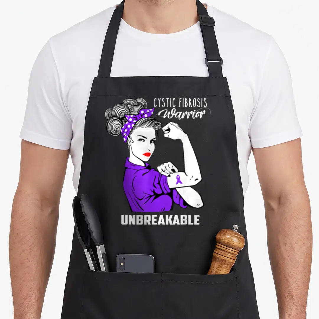 Warrior cystic fibrosis awareness apron - Cartoon woman flexing arm with purple bandana and UNBREAKABLE text — shown on a black cotton canvas apron against a white background