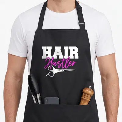 Hustler scissor hairstylist apron - White HAIR text, pink Hustler script, and white scissors graphic — shown on a black cotton canvas apron against a white background