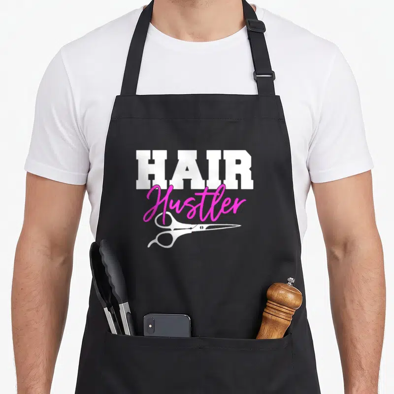 Hustler scissor hairstylist apron - White HAIR text, pink Hustler script, and white scissors graphic — shown on a black cotton canvas apron against a white background