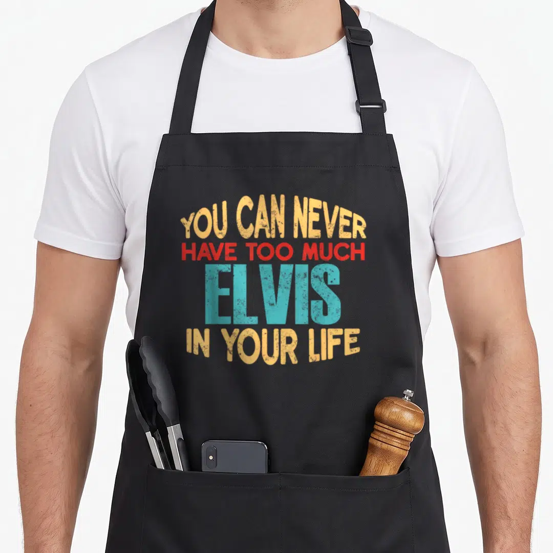 Elvis joke apron - Stacked orange red and teal text reading You Can Never Have Too Much Elvis In Your Life — shown on a black cotton canvas apron against a white background
