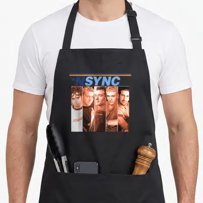 Nsync fandom apron - Five vertical photo strips of men under blue and orange bar with *NSYNC text — shown on a black cotton canvas apron against a white background