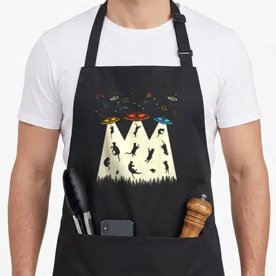 Alien kitty cat apron - Three UFOs lifting grey cat silhouettes from a forest with space objects in a cartoon style — shown on a black cotton canvas apron against a white background