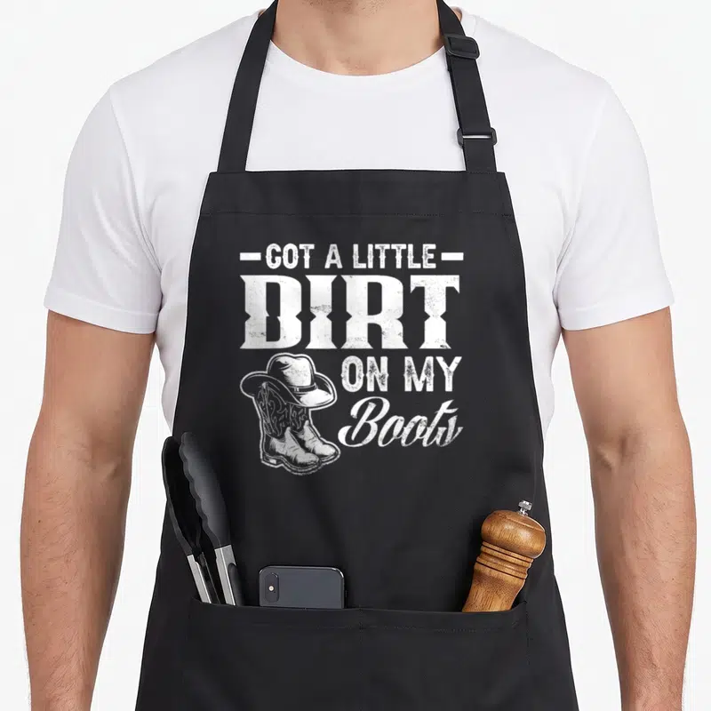 Dirt cook's country aprons - White distressed text reading Got A Little Dirt On My Boots next to cowboy boots and hat — shown on a black cotton canvas apron against a white background