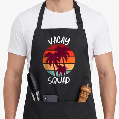 Vacay tree apron - VACAY SQUAD text above and below striped sun with three dark palm tree silhouettes in retro colors — shown on a black cotton canvas apron against a white background