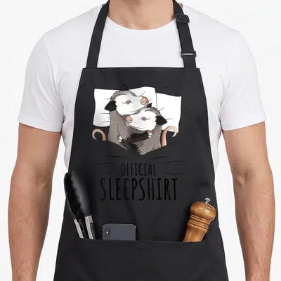 Opossum novelty apron - Two cartoon opossums cuddling under a grey blanket with OFFICIAL SLEEPSHIRT text below — shown on a black cotton canvas apron against a white background
