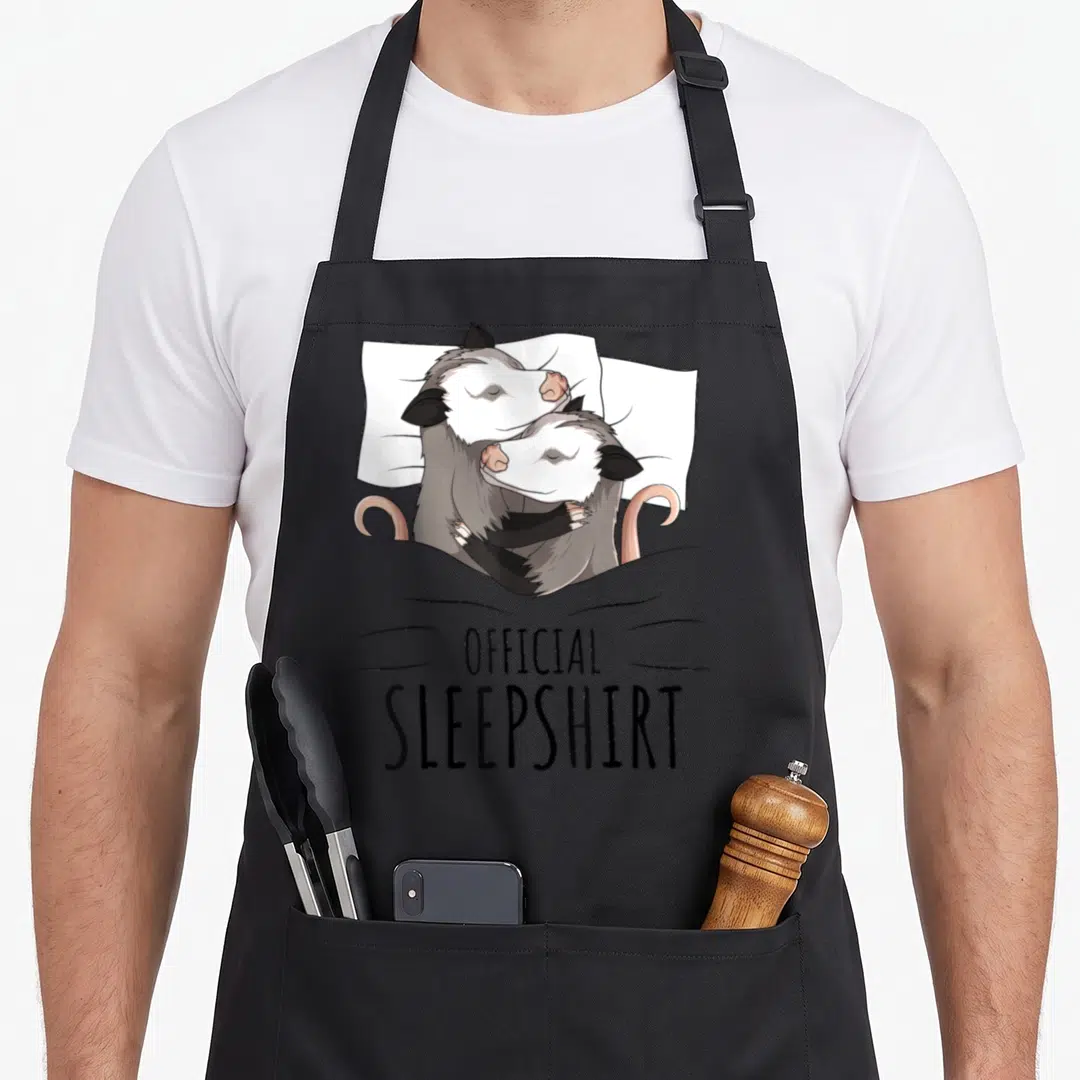 Opossum novelty apron - Two cartoon opossums cuddling under a grey blanket with OFFICIAL SLEEPSHIRT text below — shown on a black cotton canvas apron against a white background