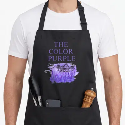 Movie lavender apron - Stacked THE COLOR PURPLE text above watercolor house and lavender field — shown on a black cotton canvas apron against a white background