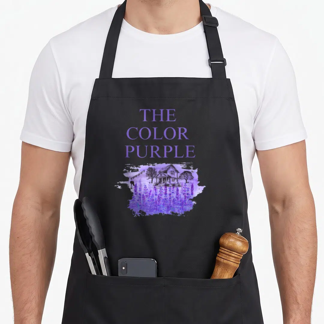 Movie lavender apron - Stacked THE COLOR PURPLE text above watercolor house and lavender field — shown on a black cotton canvas apron against a white background