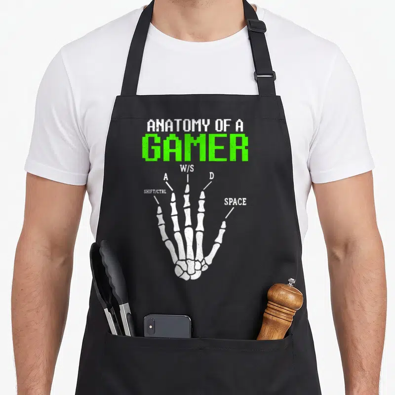 Anatomy gamer skeleton apron - White skeletal hand diagram labeled with W A S D and space bar keys in distressed green — shown on a black cotton canvas apron against a white background