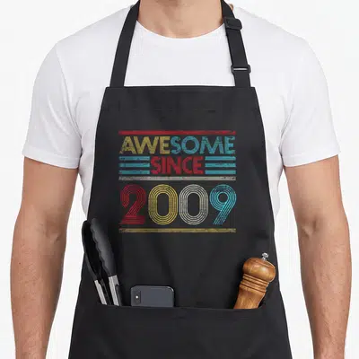 Vintage 14th birthday apron - Retro typography reading AWESOME SINCE 2009 with yellow red blue and white horizontal — shown on a black cotton canvas apron against a white background