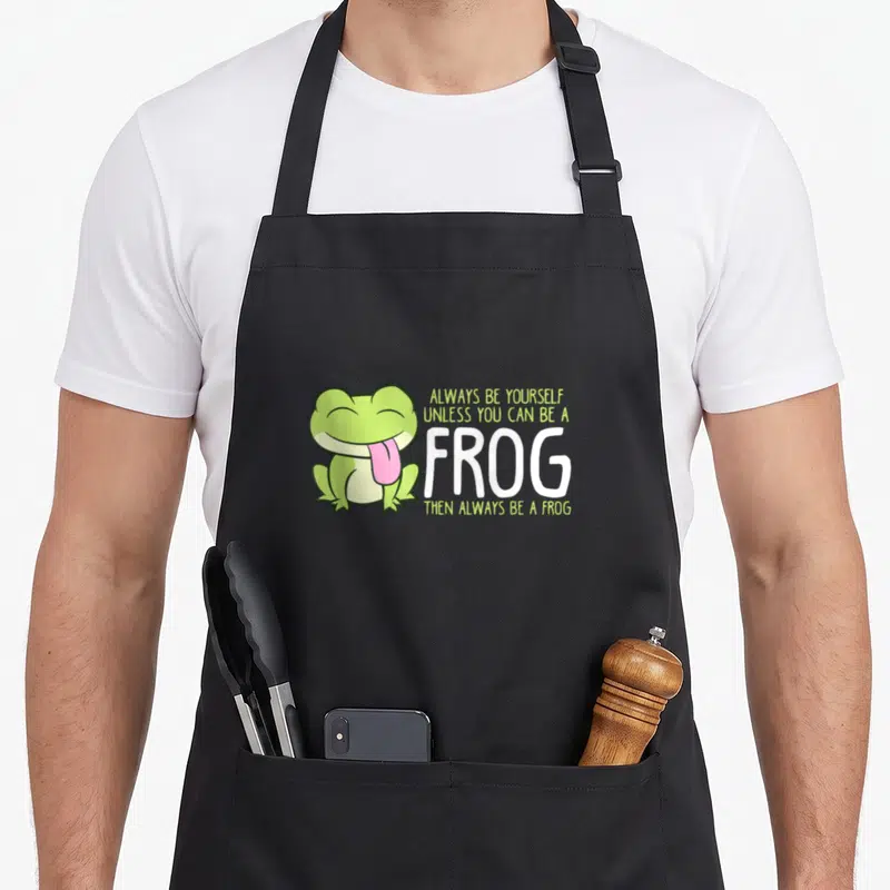 Alway frog aprons - Green cartoon frog with pink tongue and stacked white text reading ALWAYS BE YOURSELF UNLESS YOU — shown on a black cotton canvas apron against a white background
