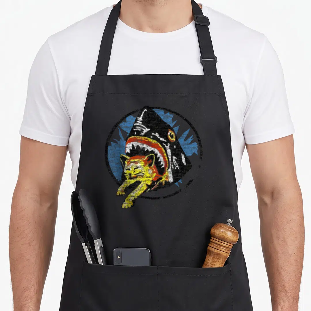 Shark cat apron - Cartoon black shark biting yellow cat inside black circle with blue splash background — shown on a black cotton canvas apron against a white background