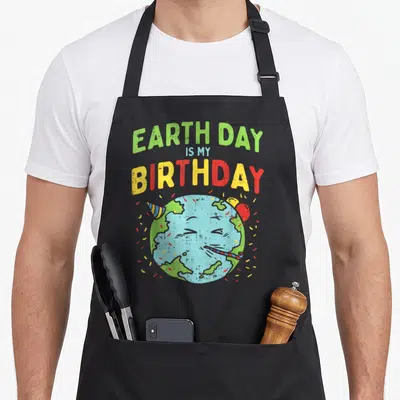 Birthday pro apron - Cartoon Earth character with party hat and blower under stacked colorful text — shown on a black cotton canvas apron against a white background