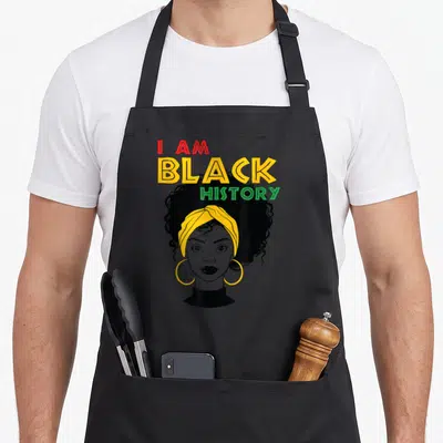 History afro african american aprons - I AM BLACK HISTORY text above woman with yellow headwrap and hoop earrings — shown on a black cotton canvas apron against a white background