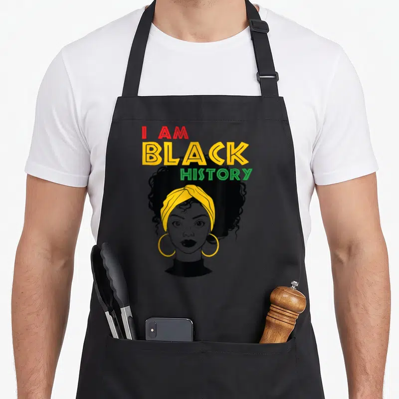 History afro african american aprons - I AM BLACK HISTORY text above woman with yellow headwrap and hoop earrings — shown on a black cotton canvas apron against a white background