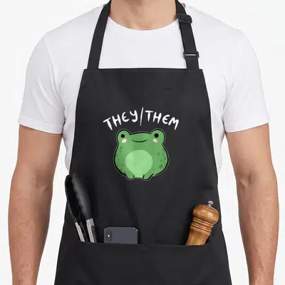 They frog aprons - Green cartoon frog with pink cheeks and white they/them text arched above — shown on a black cotton canvas apron against a white background