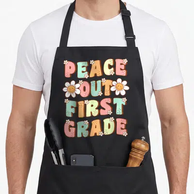 Peace retro daisy apron - PEACE OUT FIRST GRADE text with smiling white daisies in pink teal orange yellow and white — shown on a black cotton canvas apron against a white background