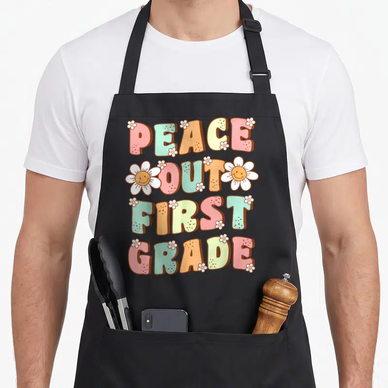 Peace retro daisy apron - PEACE OUT FIRST GRADE text with smiling white daisies in pink teal orange yellow and white — shown on a black cotton canvas apron against a white background