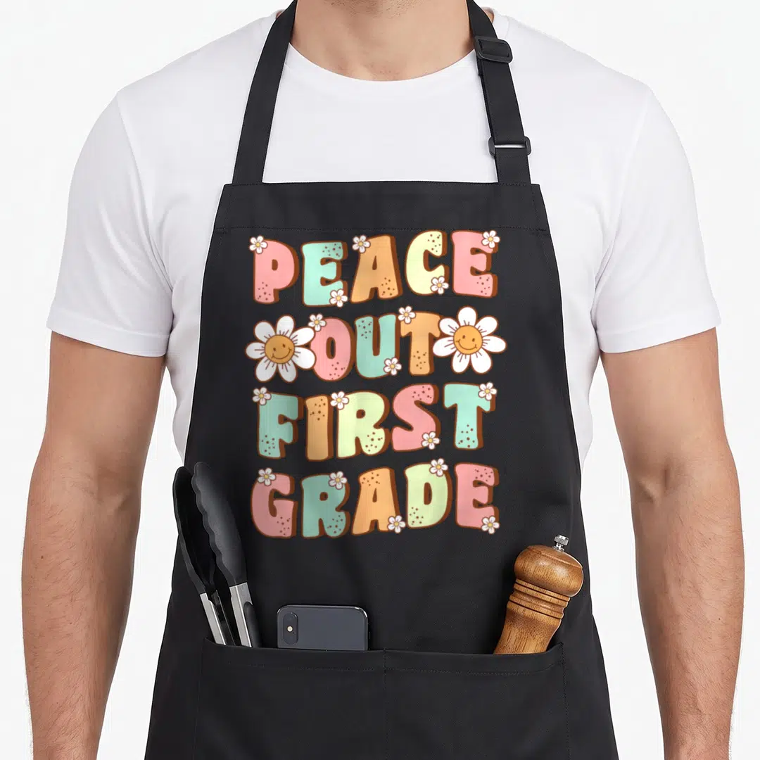 Peace retro daisy apron - PEACE OUT FIRST GRADE text with smiling white daisies in pink teal orange yellow and white — shown on a black cotton canvas apron against a white background