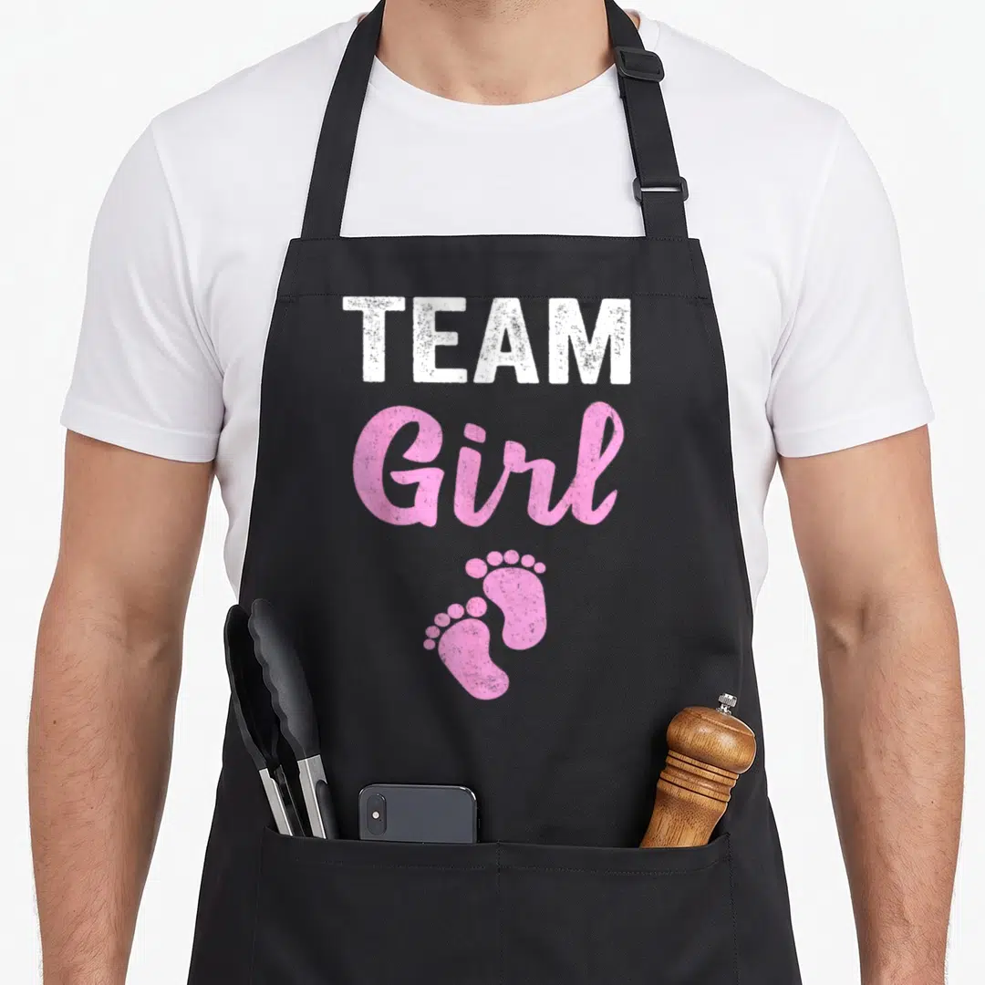 Reveal baby shower apron - Stacked white TEAM and pink Girl text above pink baby footprints on black fabric — shown on a black cotton canvas apron against a white background