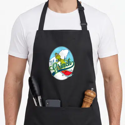 Funny snowboard holiday apron - Green Grinch character snowboarding on red board inside light blue oval with white — shown on a black cotton canvas apron against a white background