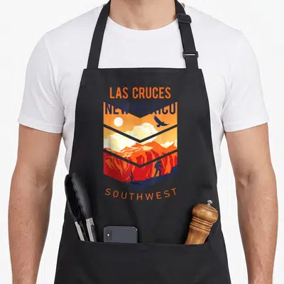 Native mexico apron - Las Cruces New Mexico Southwest shield landscape with hiker and cactus in orange and blue — shown on a black cotton canvas apron against a white background
