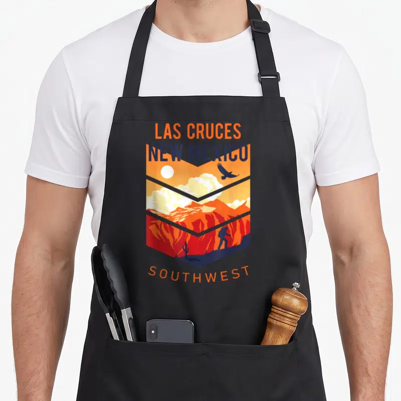 Native mexico apron - Las Cruces New Mexico Southwest shield landscape with hiker and cactus in orange and blue — shown on a black cotton canvas apron against a white background