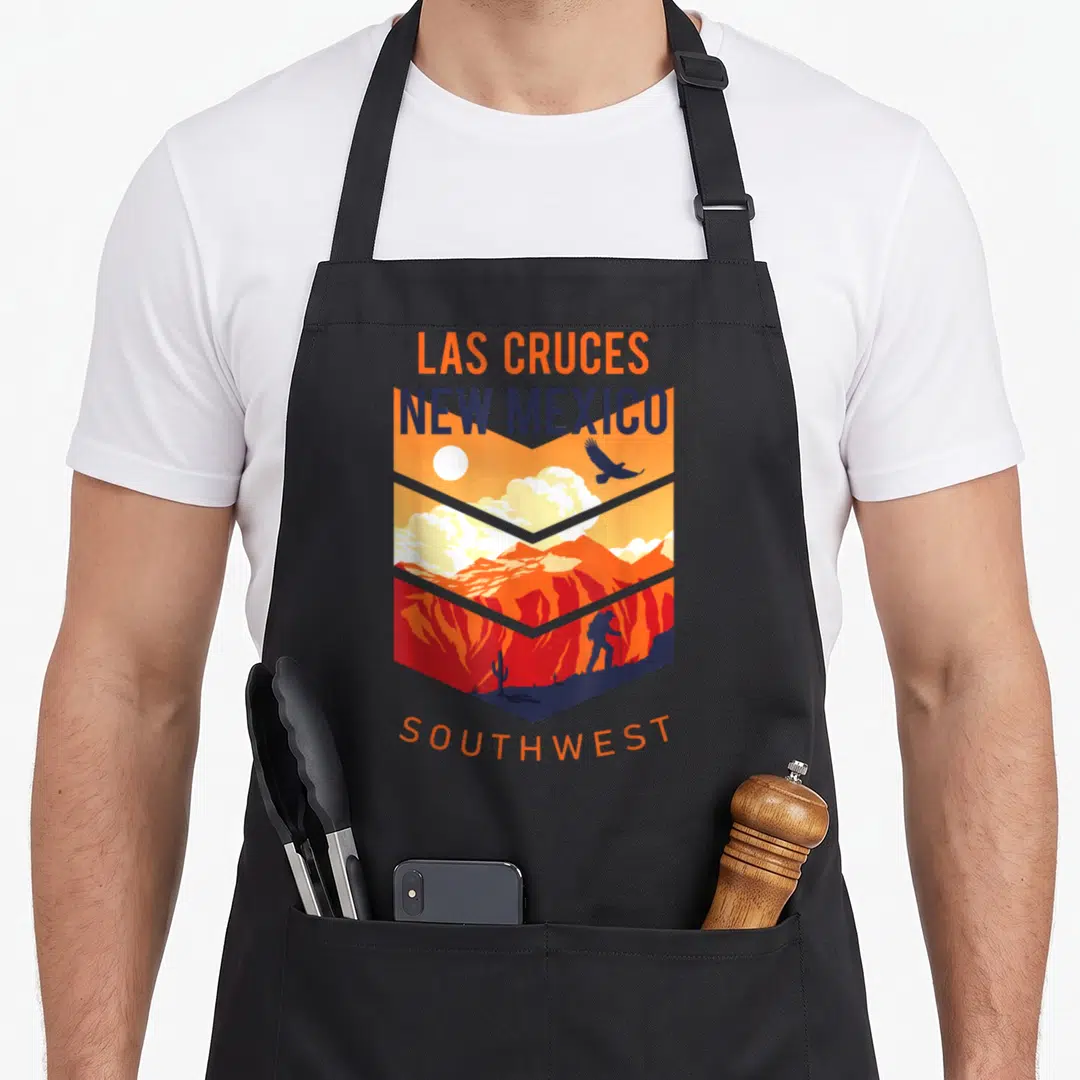 Native mexico apron - Las Cruces New Mexico Southwest shield landscape with hiker and cactus in orange and blue — shown on a black cotton canvas apron against a white background