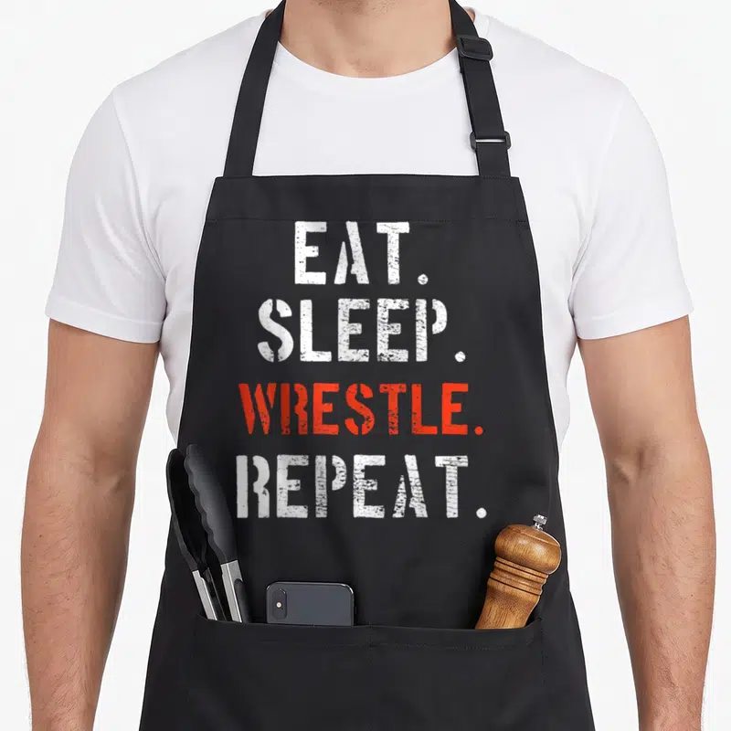 Cool wrestler wrestling apron - Stacked EAT. SLEEP. WRESTLE. REPEAT. text in white and red distressed typography — shown on a black cotton canvas apron against a white background